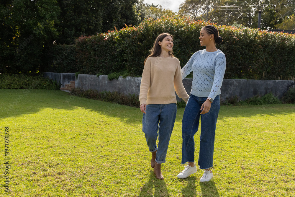 Obraz premium Diverse female friends walking together on grassy lawn near wall, wearing sweaters and jeans