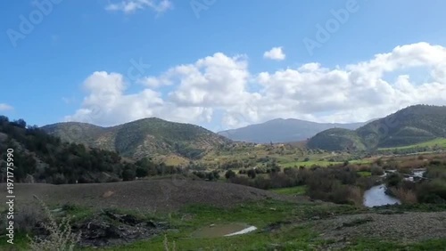 A serene video showcasing a small river flowing through a green valley with mountains in the background under a blue sky with clouds