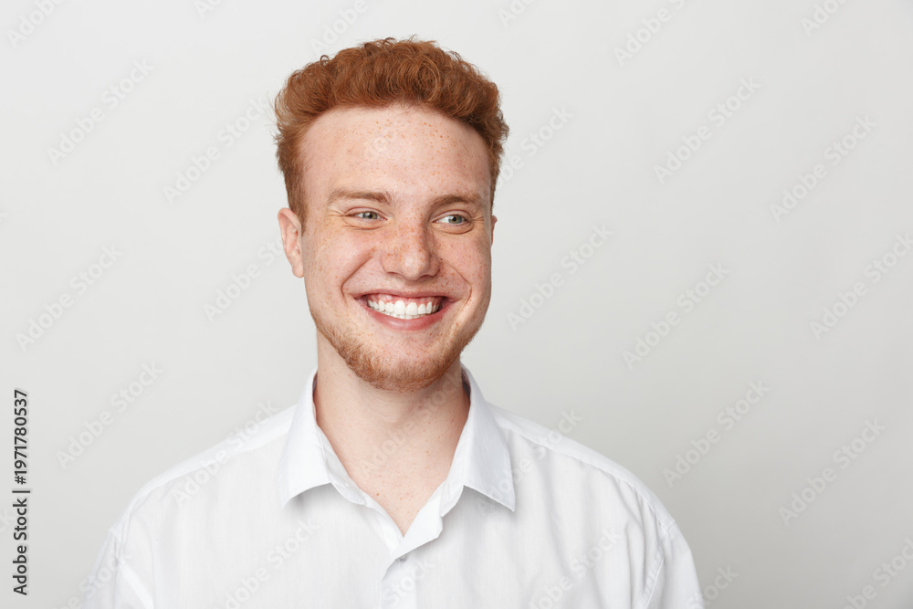 Obraz premium Smiling young man with red hair and beard wearing white shirt looks away on light background. Portrait of happy male model with natural expression and casual style in studio lighting.