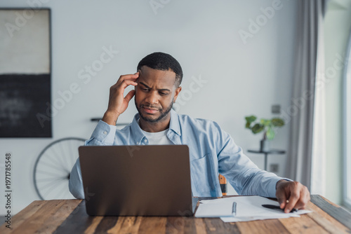 Billede på lærred A young man sits at a wooden table in a bright home office, working on a laptop and reviewing papers with a puzzled expression, suggesting concentration, remote work and problem solving