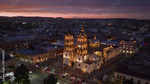 Aerial hyperlapse captures stunning Durango Cathedral bathed in golden lights at twilight in Durango, Mexico. Vibrant pink and purple sunset sky stretches across horizon as city lights twinkle below.