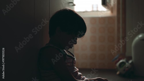 Young boy leaning against kitchen cabinet sitting on floor staring forward in silence after crying expressing childhood depression emotional exhaustion and mental health struggle