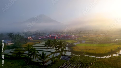 A breathtaking sunrise view of hot air balloons being prepared for launch in a mist-covered rural landscape. Surrounded by reflective rice terraces, palm trees, and a distant mountain silhouette.