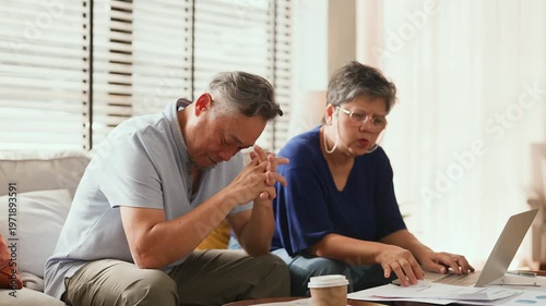 Unhappy Asian senior couple sitting on sofa at home facing financial problem, stress from debt and bills, worried about insurance and money, retirement struggle, emotional tension and family hardship.
