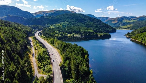 Aerial view of a scenic mountain highway winding beside a large blue lake, lush green forest landscape, travel and adventure destination photography