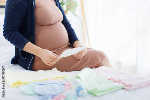 Asian young pregnant woman sitting relax on white bed in bedroom at home waiting and preparing baby clothes accessory baby shower infant care children products love healthy happy pregnancy motherhood.