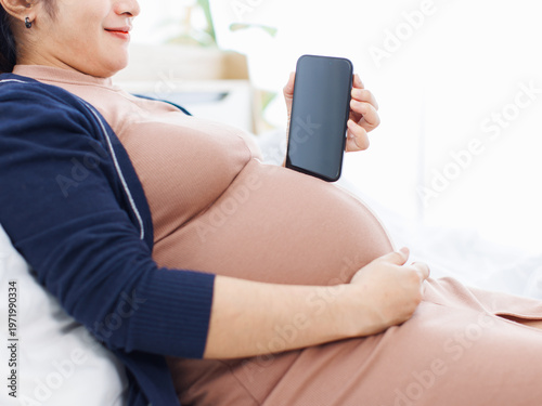 Asian happy young calm pregnant woman sitting on white bed in bedroom with mobile smartphone reading research info prenatal health shopping online relaxed maternity love wellness pregnancy motherhood.