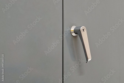 Foto Glossy silver metal cabinet door handle with built-in lock cylinder, mounted on a flat grey metal cabinet door
