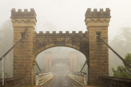 An historic single span suspension bridge called Hampden Bridge opened in 1898 with crenellated sandstone turrets and cables in an early morning mist in Kangaroo Valley in New South Wales, Australia.