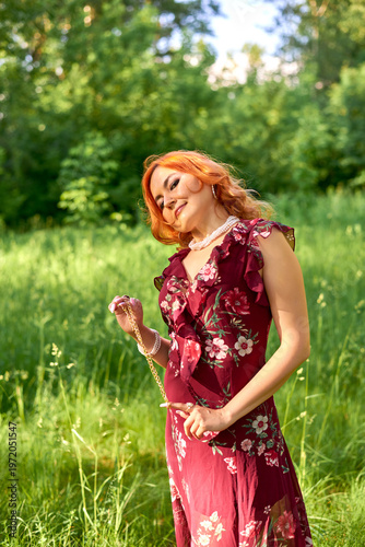A woman with red hair is standing in a sunlit meadow. She is wearing a floral-print dress and is looking at the camera. She is surrounded by green grass and trees. This is perfect for a lifestyle and 
