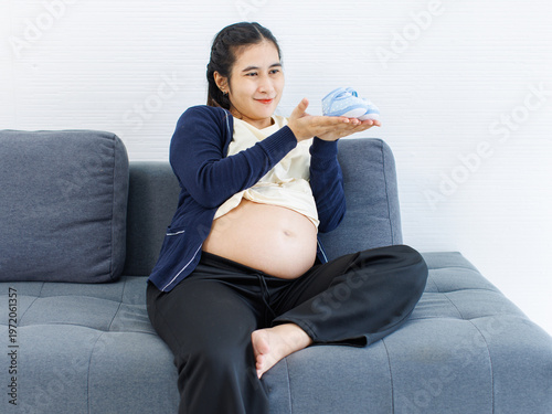 Asian young pregnant woman sitting smile on sofa happy holding cute baby shoes on big belly tummy get ready mother care love healthy pregnancy motherhood maternity expecting newborn extending family.