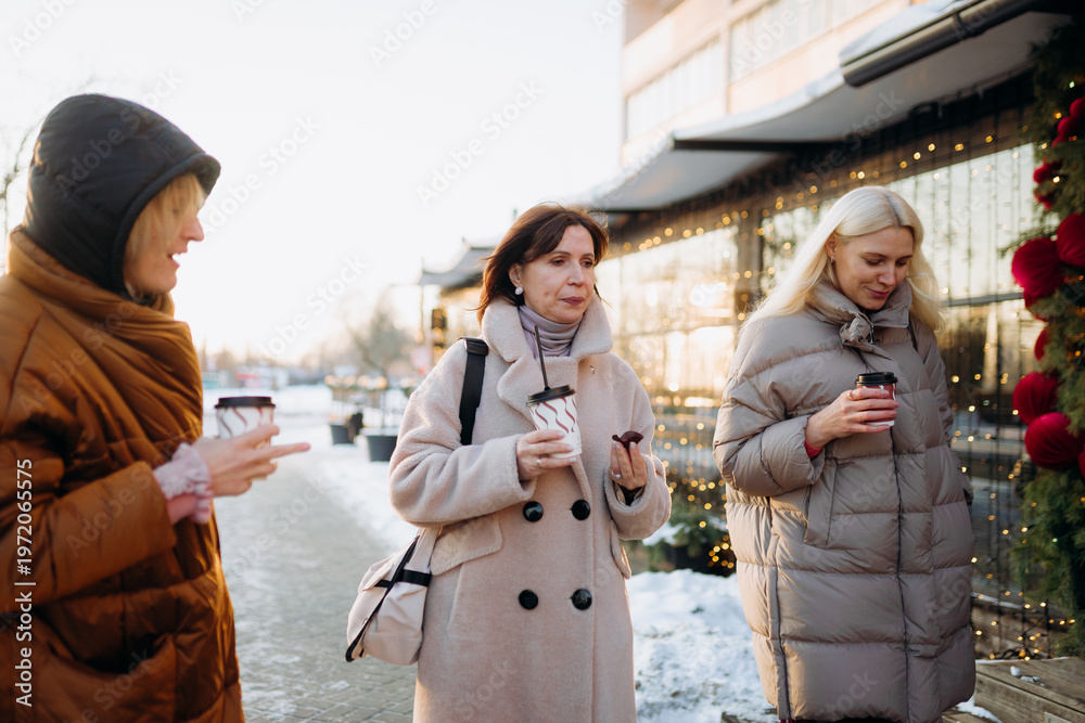 Obraz premium Three friends stroll down a snowy street, each holding a warm beverage. They share smiles and laughter, enjoying the winter sunlight and festive decorations around them.