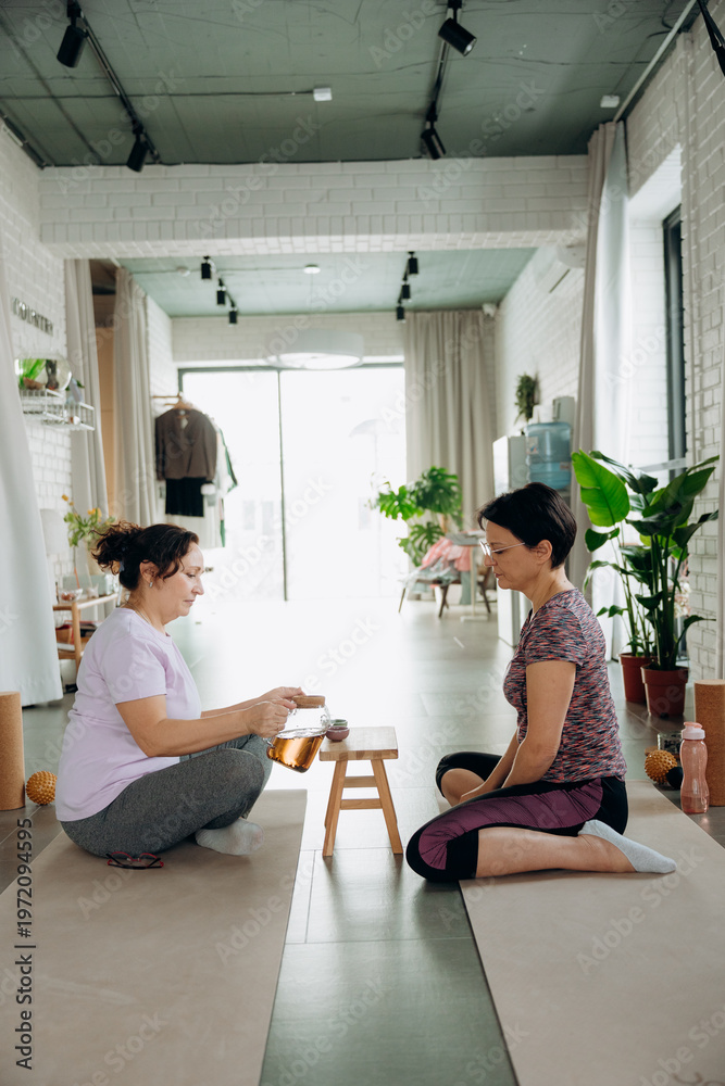 Fototapeta premium Two women sit cross-legged on soft mats, focusing on a small table with calming items. Natural light fills the peaceful studio, creating a relaxing atmosphere for their meditation.