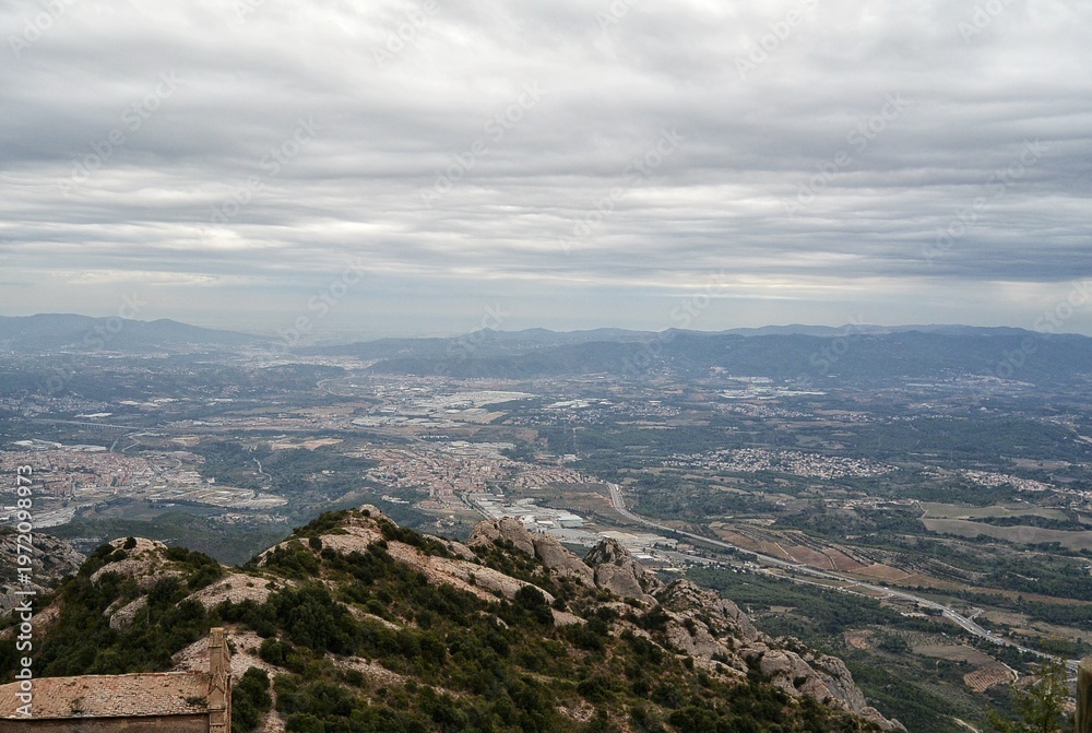 Obraz premium A high angle panoramic view from Montserrat mountain in Catalonia, Spain. Rocky peaks in the foreground, green valley with towns and roads under a grey cloudy sky during a daytime in the summer