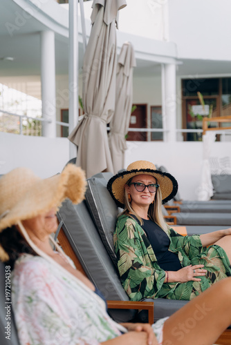 Foto Two women sit in lounge chairs at a resort pool, wearing large hats and casual outfits