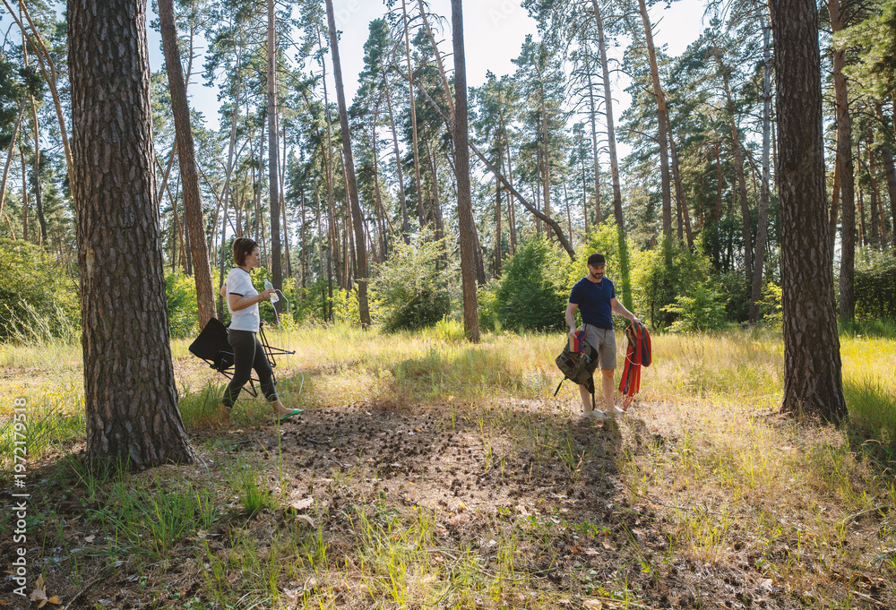 Fototapeta premium Young Couple Setting Up Camp in Sunny Pine Forest with Hammock and Chair