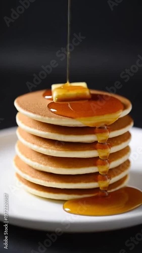 Close-up shot of a stack of six fluffy pancakes on a white plate with a pat of butter on top while amber maple syrup is being poured over the side against a dark background.