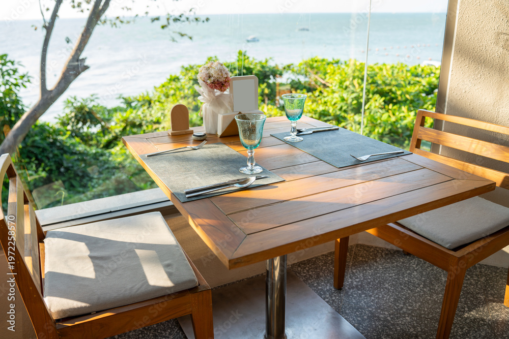 Naklejka premium Table set for two with blue water glasses and silver cutlery on a wooden patio overlooking the ocean. Ideal for luxury seaside dining, beach cafe, and summer vacation lifestyle themes.