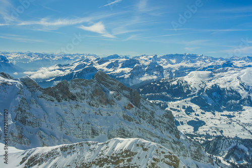 Snow covered mountain range under clear blue sky, showcasing rugged peaks and expansive valleys. serene landscape captures beauty of winter in mountains, with distant clouds adding depth
