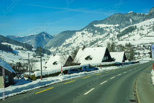 Snow covered alpine village with traditional houses and winding road under clear blue sky, surrounded by majestic mountains. serene winter landscape evokes sense of tranquility