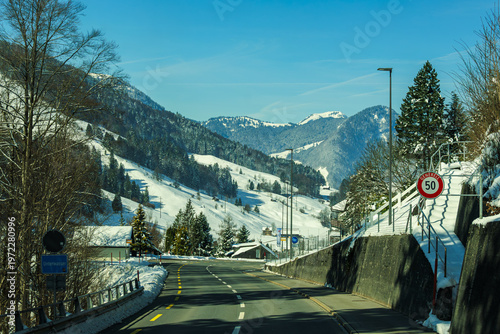 Snowy mountain road with clear blue sky, surrounded by trees and hills, creating serene winter landscape. speed limit sign is visible, indicating 50 km/h