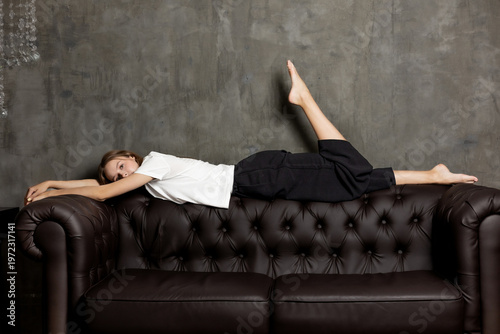 Girl lying on a brown leather couch with one leg raised and arms stretched in a modern interior setting