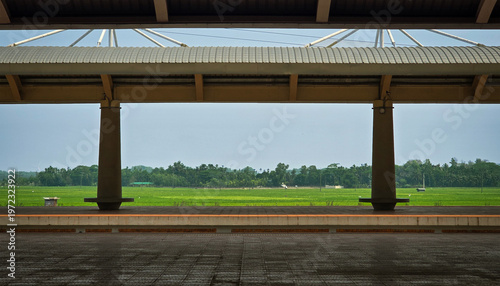 Cinematic symmetrical view of an empty railway station platform overlooking lush green rural fields under a clear sky, featuring modern architectural pillars and a wide scenic perspective.