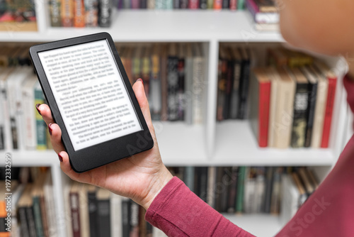 a woman holds an e-book in her hand. a woman's hand holding an e-book. A woman reads an e-reader near a bookcase. an e-book against the background of paper books. A woman reading an e-book at home