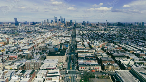 Long axis city sweep along Broad St highlights uninterrupted neighborhood grids extending toward downtown towers. Light roof membranes, muted masonry, bright midday clarity define Philly, PA
