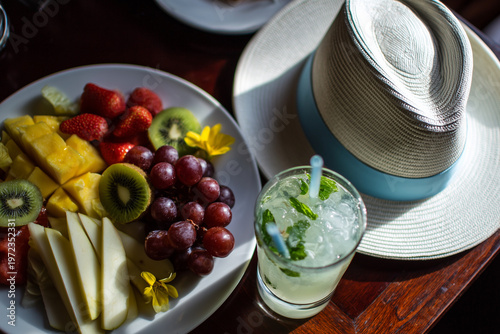 Fresh fruit, straw hat, and cold drink arranged in sunlight, evoking summer leisure, travel ease, refreshment, simplicity, and abundance.