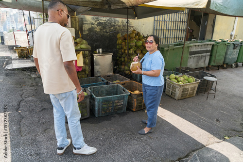 Man and woman talk at marketplace near trash bins during a sunny day while selecting fresh produce in an urban area with buildings