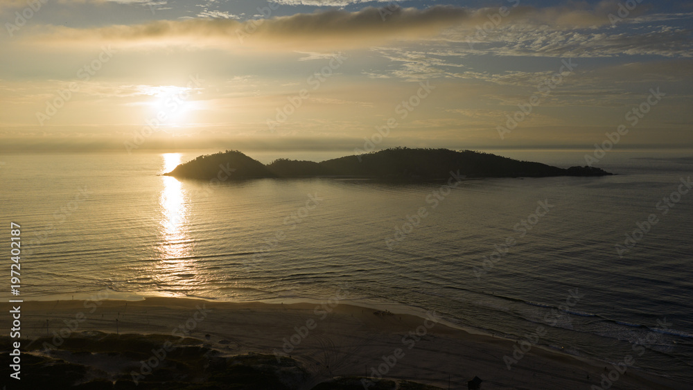 Fototapeta premium Aerial view of Campeche Island silhouette at sunrise with golden light reflecting on calm sea, Florianopolis.