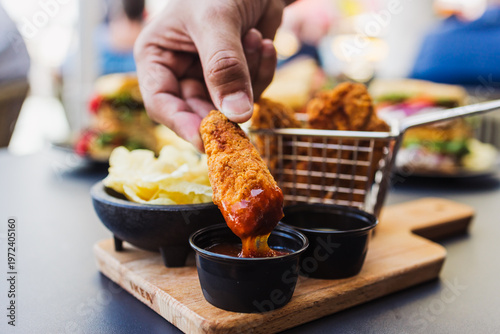 Slika na platnu Hand dipping a crispy chicken nugget into red dipping sauce on rustic wooden ser