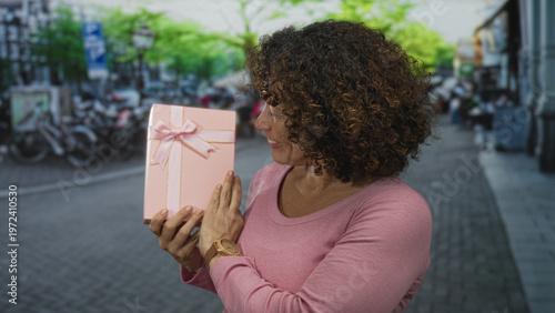 Obraz na plátně Woman smiles and holds pink giftbox on a sunny colorful leafy bustling street; gratitude celebration joy
