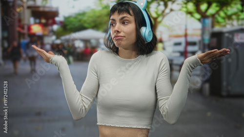 Φωτογραφία Young hispanic woman wearing blue cat ear headphones and a cropped top shrugging arms on urban street with blurred pedestrians behind; uncertainty