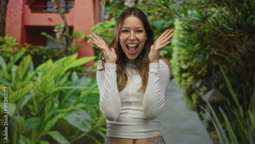 Canvas-taulu Young hispanic woman smiling and raising hands near face showing navel on a walkway by a building lined with plants; joy celebration