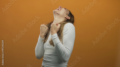 Canvastavla Young hispanic woman clenching fists and looking upward, smiling in a white top in orange studio; victory joy celebration