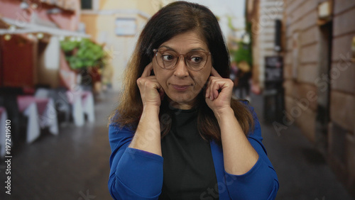 Fotografia Hispanic woman covers ears with fingers on street lined with cafe tables and potted plants; frustration