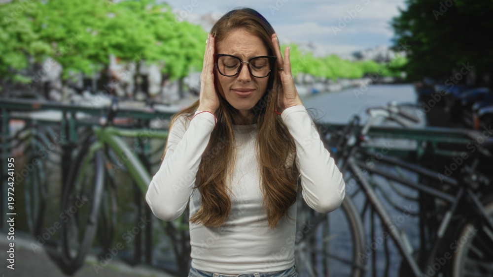Fototapeta premium Woman holding hands to her ears and wincing on an amsterdam street bridge by canal; frustration stress.