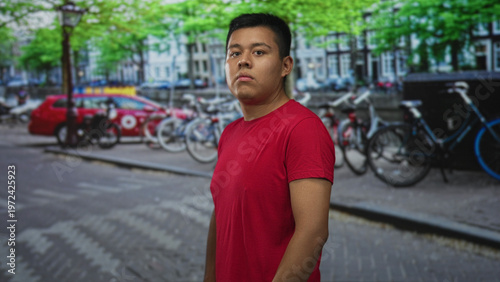 Bild auf Leinwand Man in a red t shirt standing and looking at camera on a city street, cobblestone pavement visible; quiet confidence