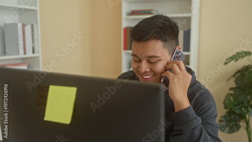Tableau sur toile Man holds smartphone to ear, smiling at computer monitor while seated at a desk in a building; contentment