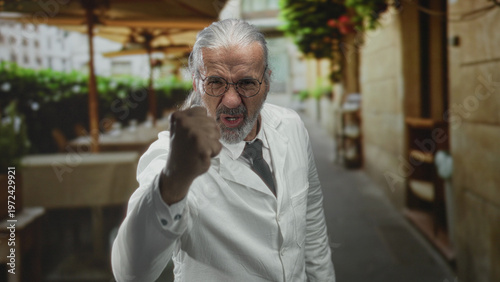 Фотография Scientist man with long grey hair and glasses raising a clenched fist toward camera on street terrace outside restaurant while wearing a white lab coat; anger defiance