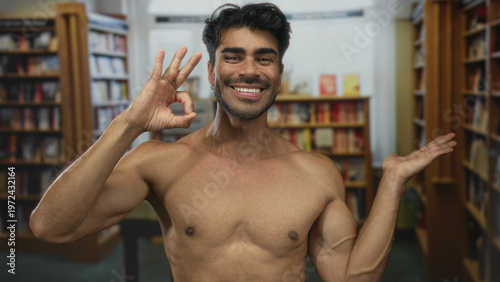 фотографія Man reveals bare chest, smiles and makes ok sign with hand in library building; approval confidence