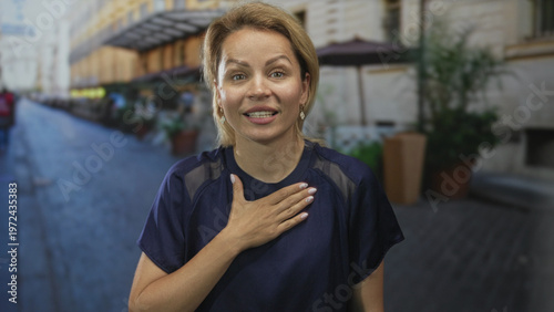 Fotografia Woman stands with hand on chest gesture on a busy cobblestone street between historic building facades; sincerity