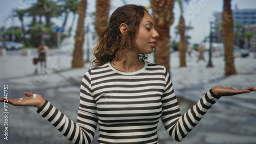 Foto Young woman holding palms up in a shrug pose on a city street wearing a black and white striped shirt; thoughtful balance