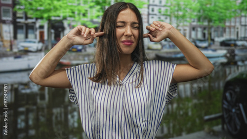 Photography Young hispanic woman in a striped shirt plugs fingers into ears on a street in amsterdam; annoyance defiance