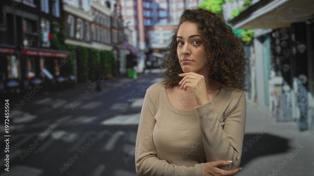 Fototapeta premium Woman with hand on chin at a city street crosswalk, neutral expression and folded arm across body, visible ring on finger; quiet contemplation.