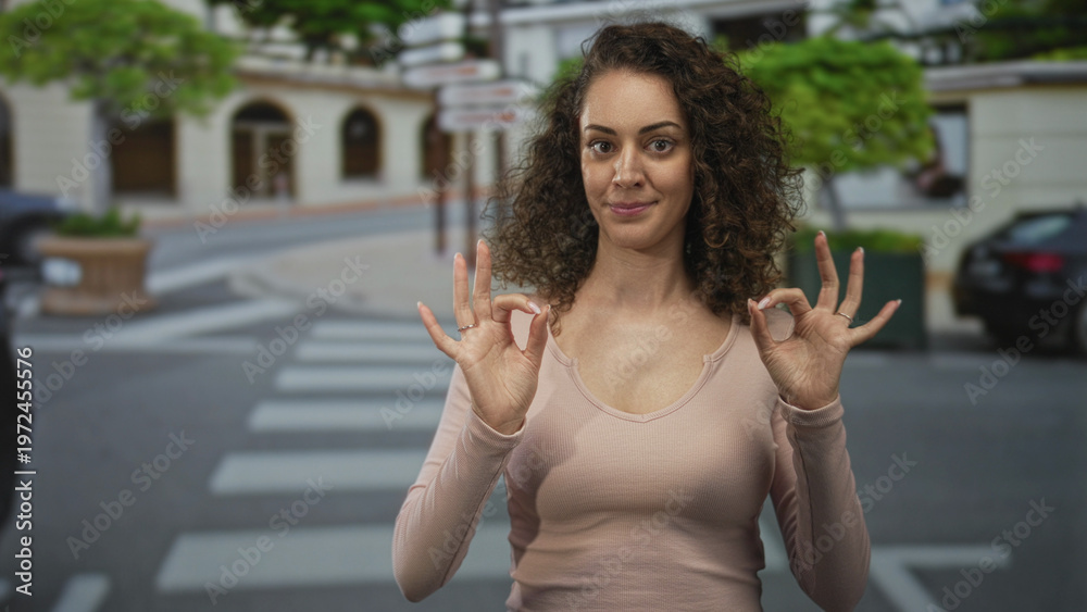 Fototapeta premium Young hispanic woman making ok sign with both hands while standing on a city crosswalk and sidewalk near parked cars and planters; confidence.