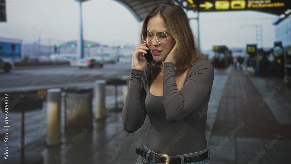 Fototapeta premium Woman holding phone to ear with hand covering ear and furrowed brow at airport curbside under departure canopy; anxiety travel delay.