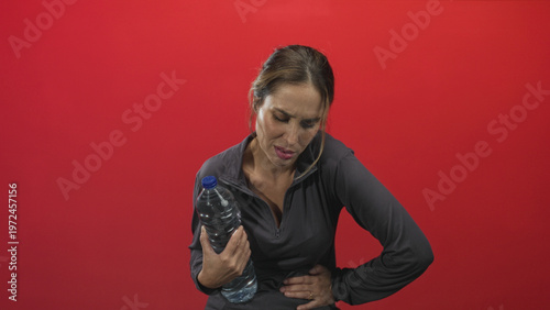 Bild auf Leinwand Woman holding water bottle with hand on stomach showing stomachache and pained expression in studio; pain concern recovery hope